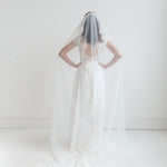 A woman in a white wedding dress stands with her hands on her hips, wearing a long white veil that extends behind her.