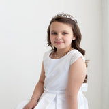 A young girl in a white dress with a tiara smiles at the camera.