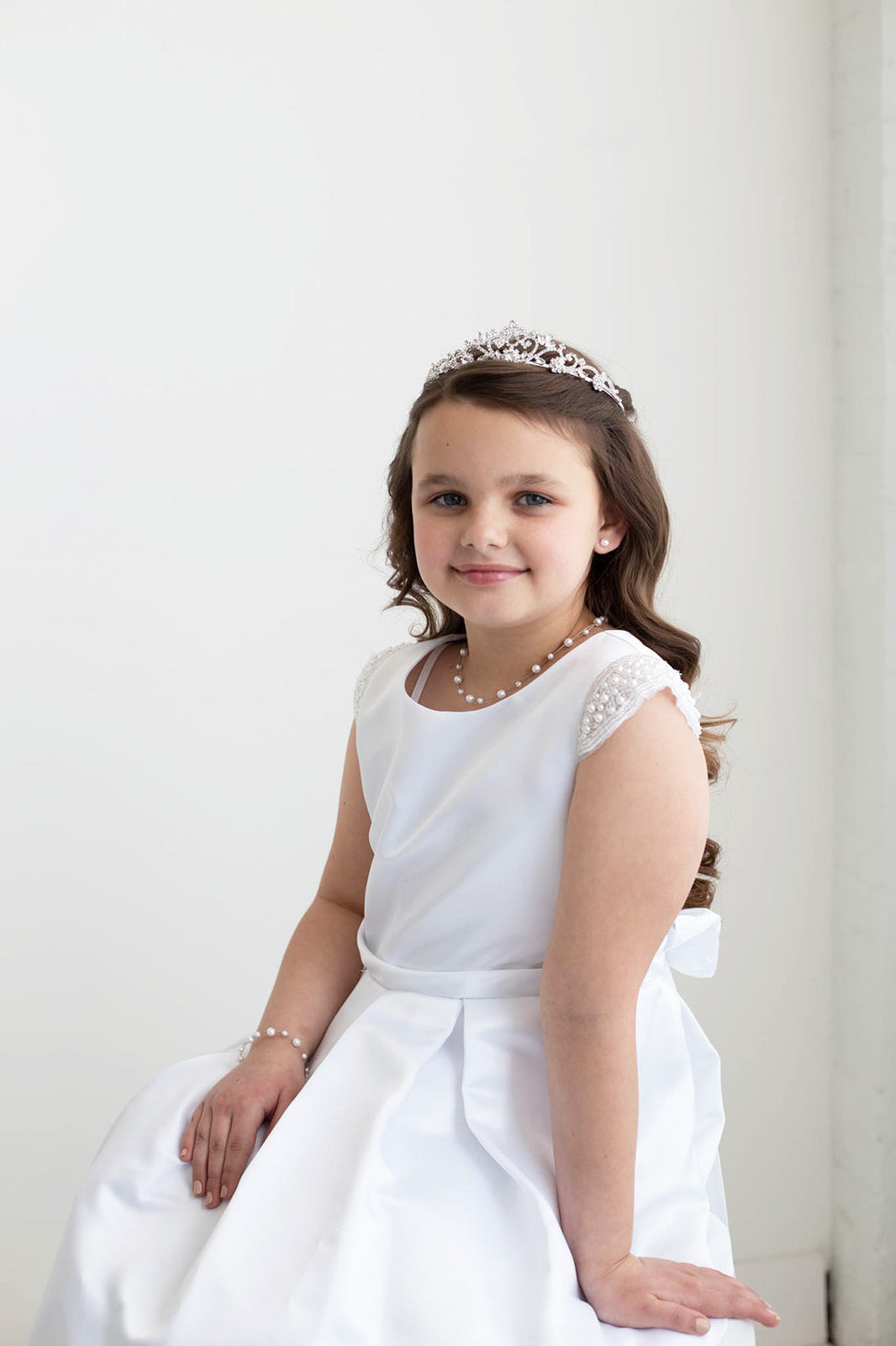 A young girl in a white dress with a tiara smiles at the camera.