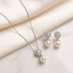 A delicate necklace with a pendant and earrings made of pearls and diamonds are displayed on a white fabric surface.