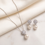 A delicate necklace with a pendant and earrings made of pearls and diamonds are displayed on a white fabric surface.