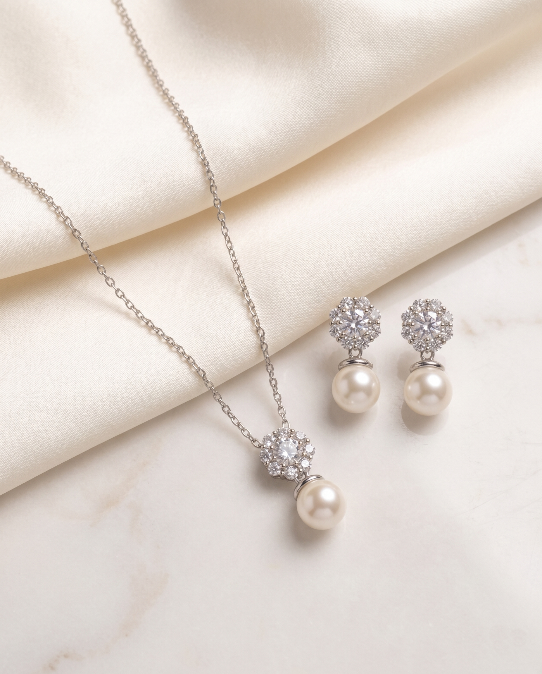 A delicate necklace with a pendant and earrings made of pearls and diamonds are displayed on a white fabric surface.