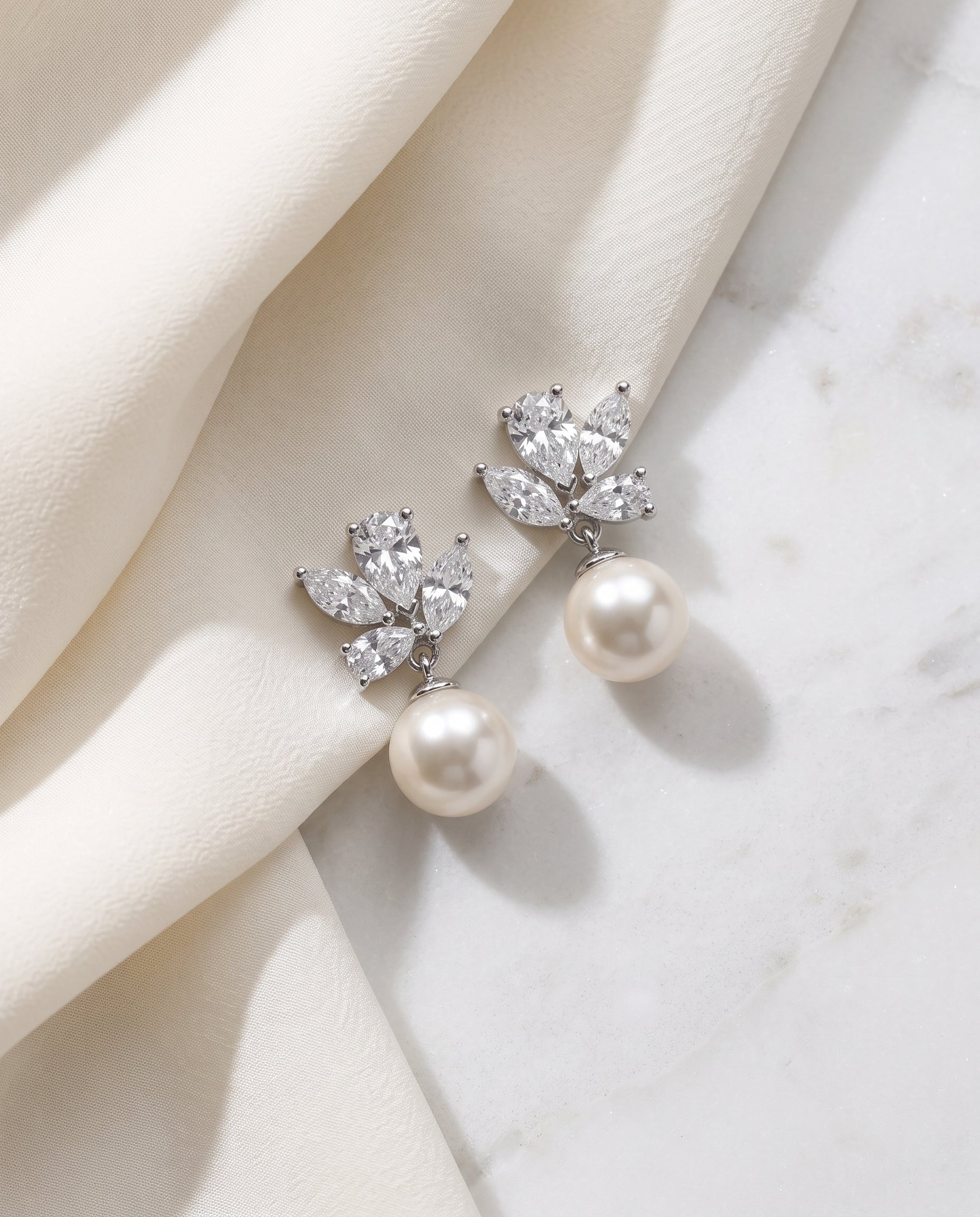 A pair of elegant earrings with a pearl and crystal embellishment, resting on a white fabric surface.