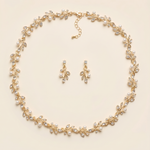 A delicate gold necklace with a leaf-shaped pendant and a pair of dangling earrings are arranged in a circular pattern on a light-colored background.