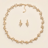 A delicate gold necklace with a leaf-shaped pendant and a pair of dangling earrings are arranged in a circular pattern on a light-colored background.