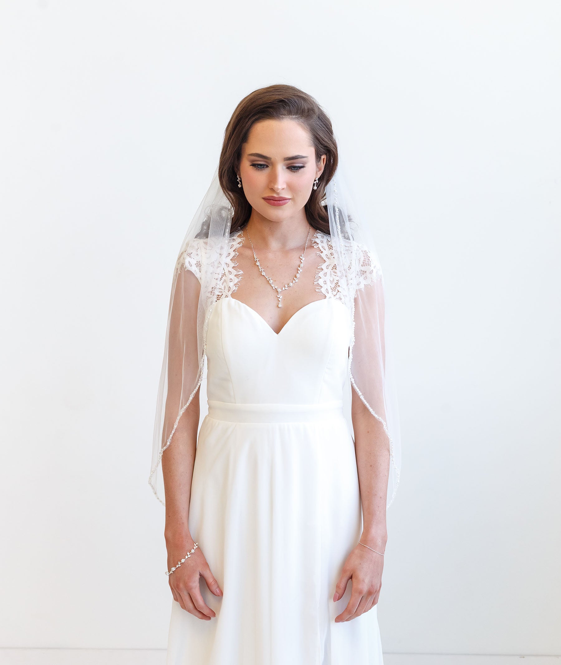 A woman in a white wedding dress with a veil stands against a white background, looking down with a thoughtful expression.