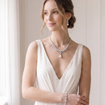 A woman in a white dress with a necklace and bracelet stands in a room with a window.