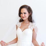 A woman in a white wedding dress with a veil stands against a white background, smiling at the camera.