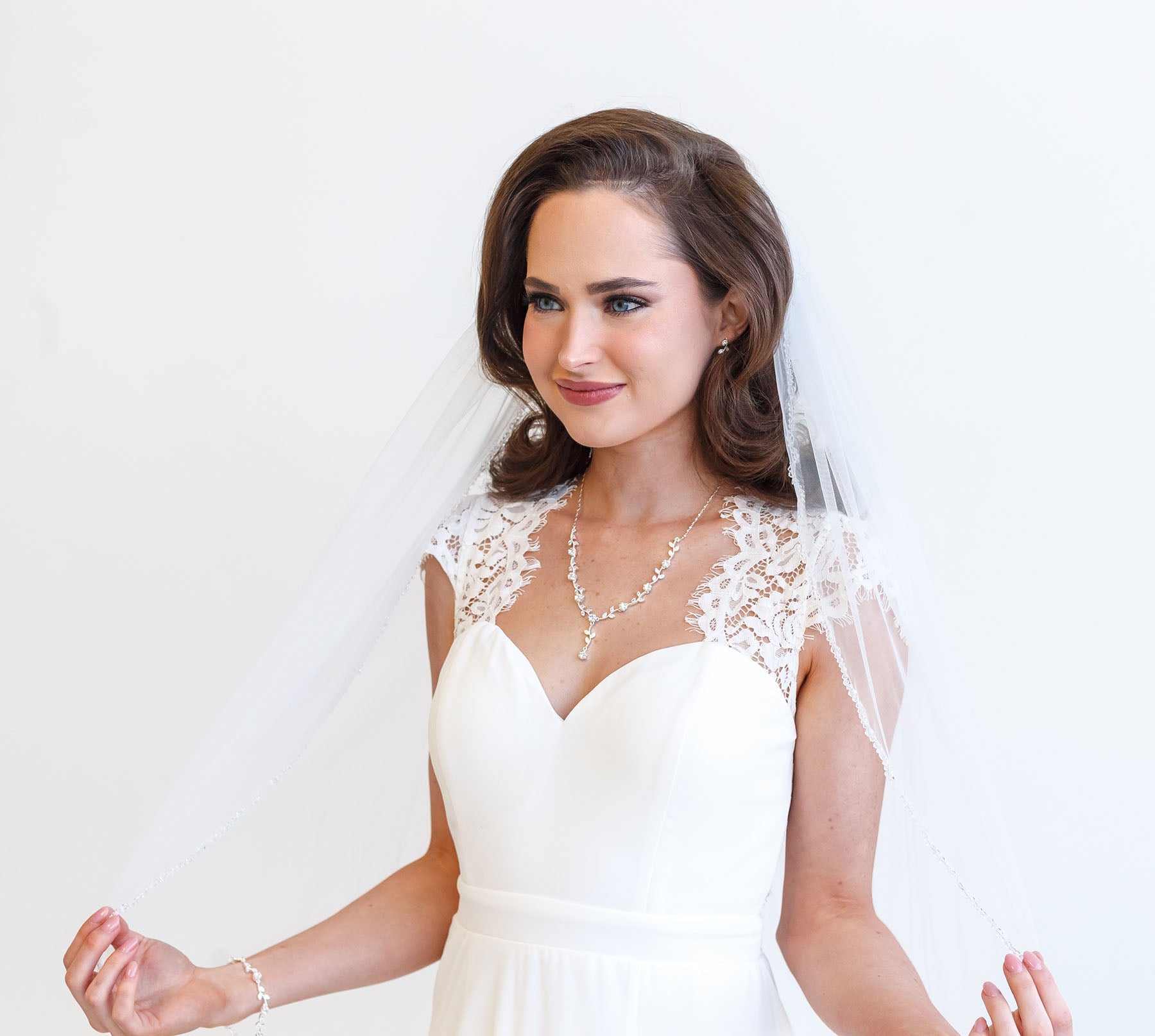 A woman in a white wedding dress with a veil stands against a white background, smiling at the camera.