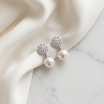 A pair of elegant earrings with white pearls and sparkling crystal accents, resting on a white fabric surface.
