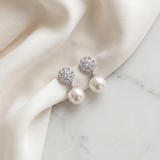 A pair of elegant earrings with white pearls and sparkling crystal accents, resting on a white fabric surface.