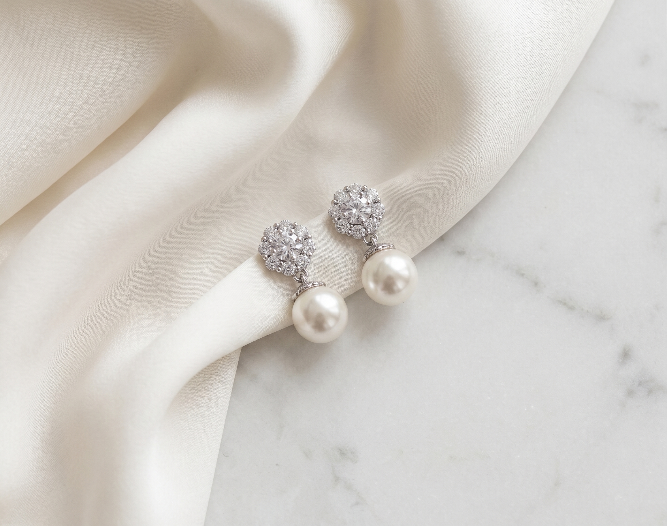 A pair of elegant earrings with white pearls and sparkling crystal accents, resting on a white fabric surface.