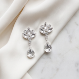 A pair of elegant diamond earrings with clear gemstones, resting on a white fabric surface.