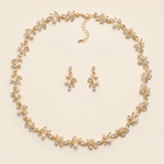 A delicate gold necklace with a leaf-shaped pendant and a pair of dangling earrings are arranged in a circular pattern on a light-colored background.