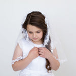 A young girl in a white dress and veil stands against a white background, her hands clasped together in front of her.