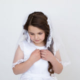 A young girl in a white dress and veil stands against a white background, her hands clasped together in front of her.