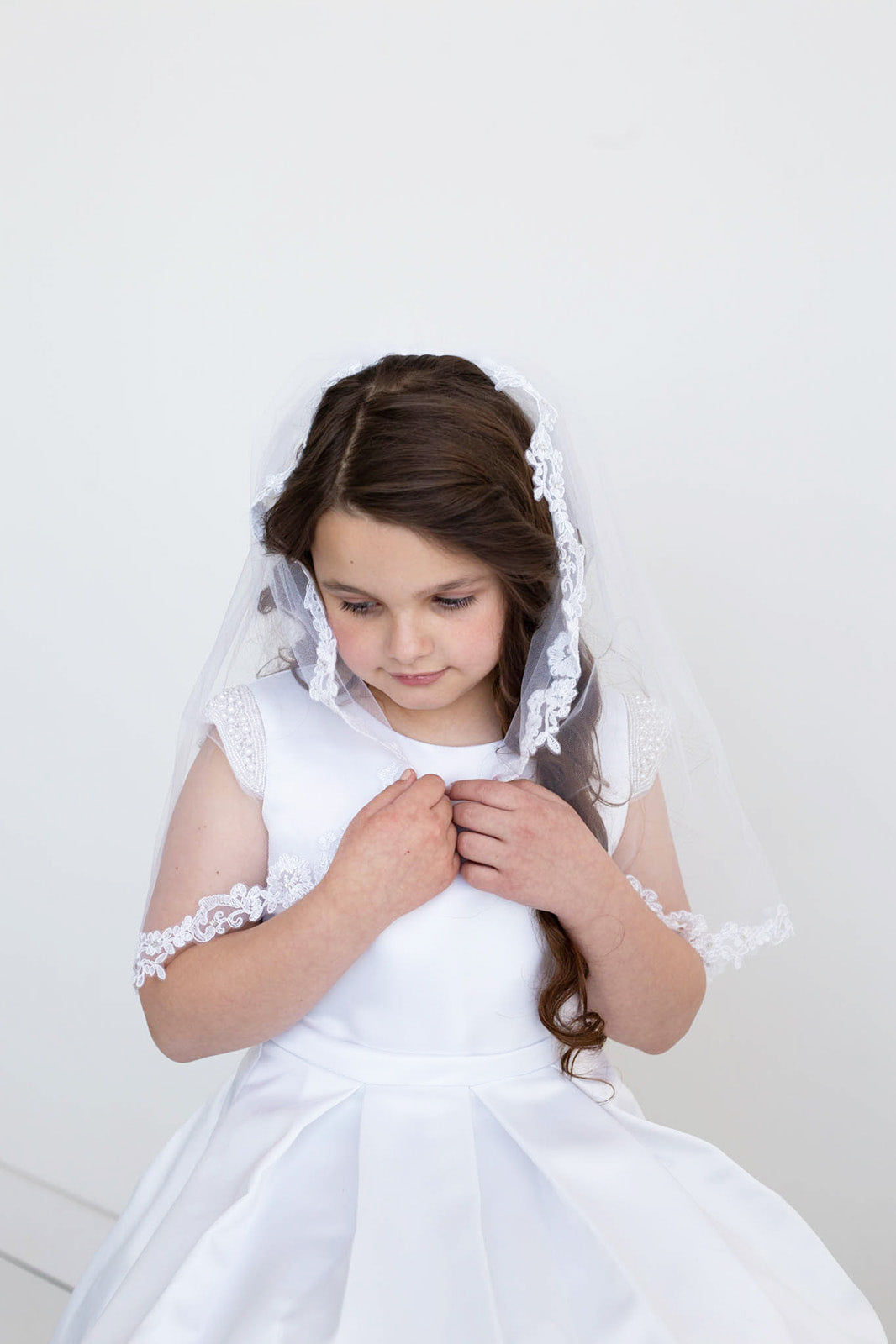A young girl in a white dress and veil stands against a white background, her hands clasped together in front of her.