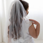 A young girl in a white dress with a veil stands in front of a white background, facing away from the camera.