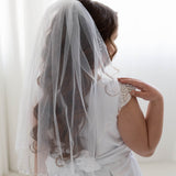 A young girl in a white dress with a veil stands in front of a white background, facing away from the camera.