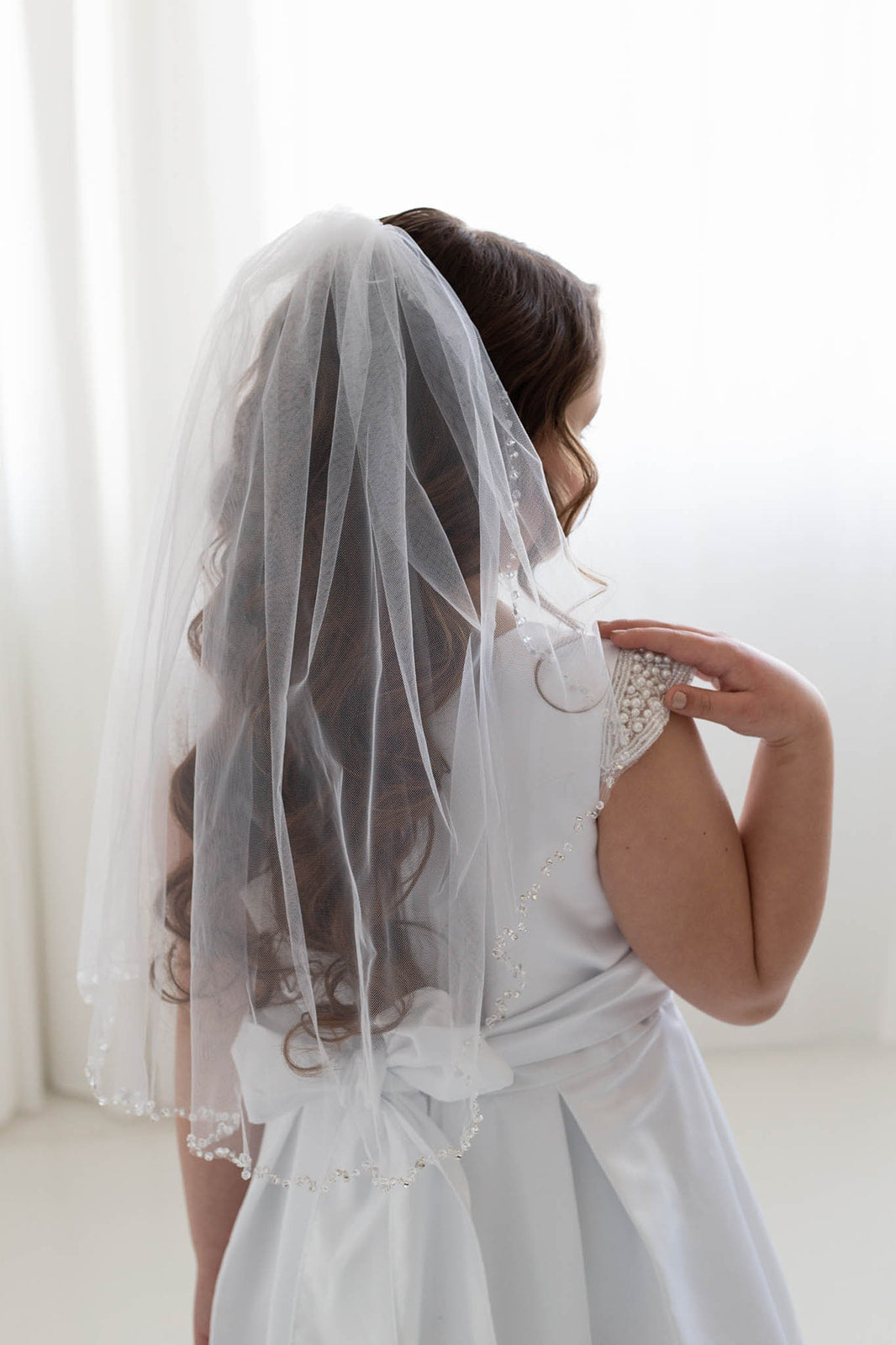 A young girl in a white dress with a veil stands in front of a white background, facing away from the camera.