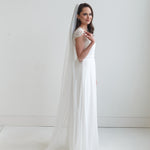 A woman in a long white wedding dress with a veil stands in a studio setting, smiling at the camera.