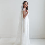 A woman in a long white wedding dress with a veil stands in a studio setting, smiling at the camera.
