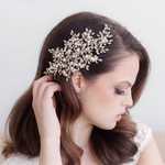 A woman with long brown hair is wearing a decorative headpiece with small white flowers or pearls. She is looking down and touching her hair.