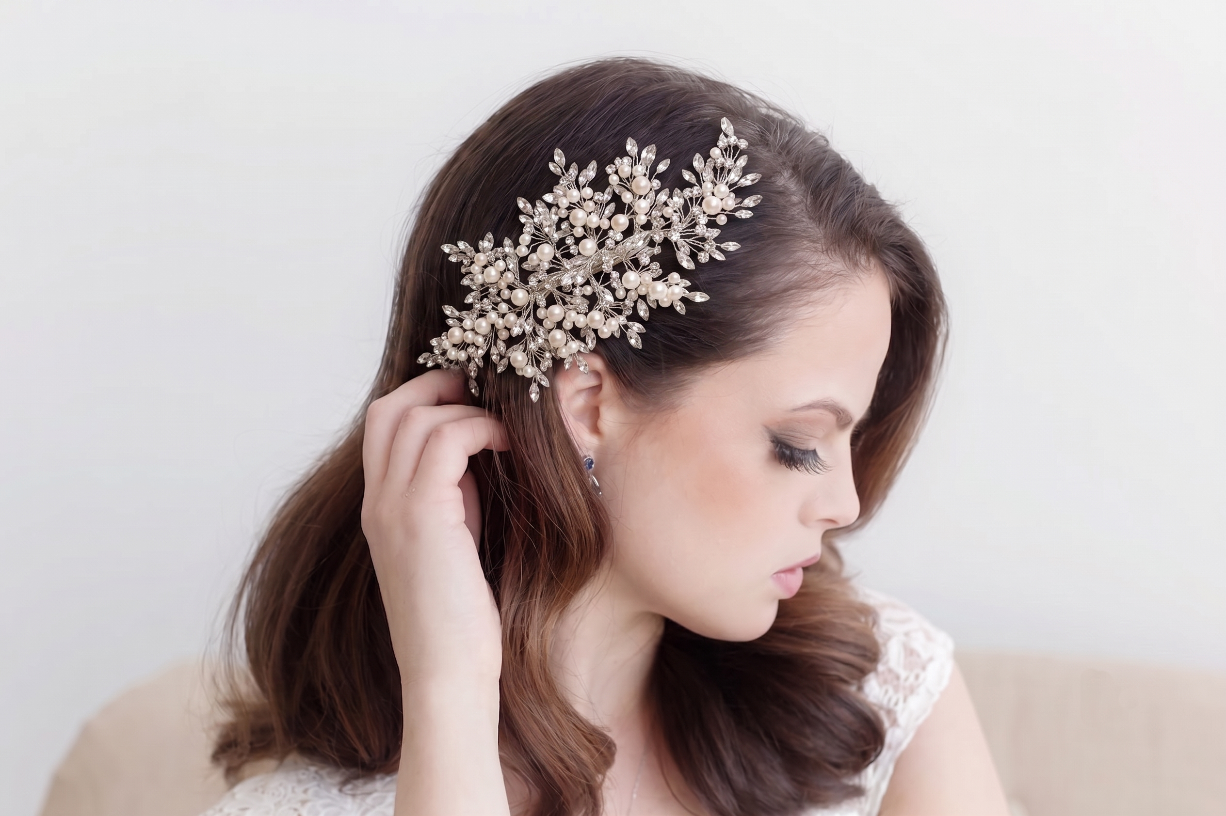 A woman with long brown hair is wearing a decorative headpiece with small white flowers or pearls. She is looking down and touching her hair.