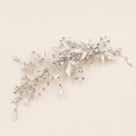 A decorative hair accessory made of silver and crystal elements, including leaves and flowers, is displayed against a light beige background.