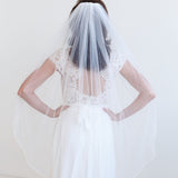 A woman wearing a white wedding dress with a long veil stands with her hands on her hips, facing away from the camera.