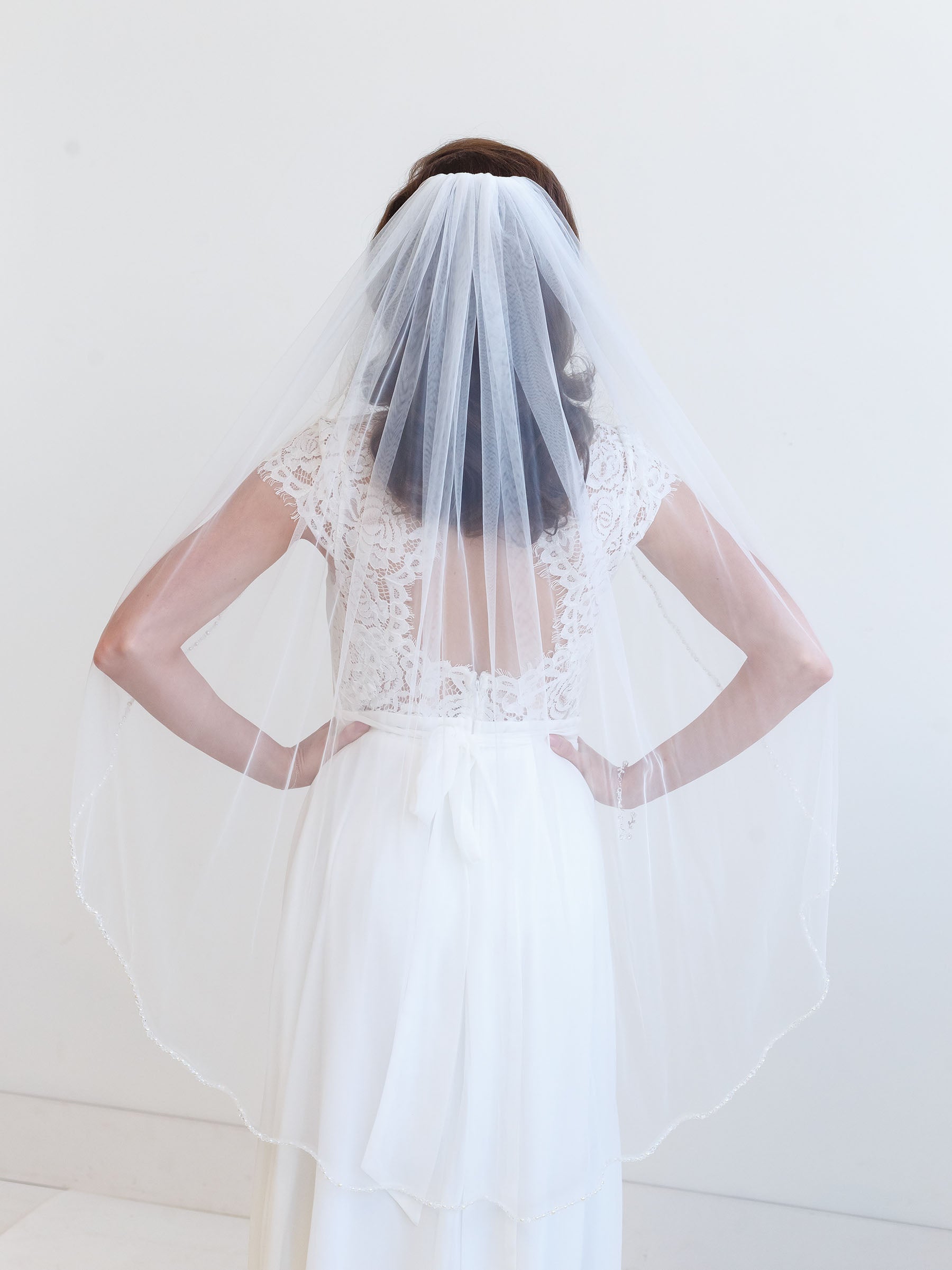 A woman wearing a white wedding dress with a long veil stands with her hands on her hips, facing away from the camera.