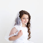 A young girl in a white dress with a veil stands against a plain white background, smiling at the camera.