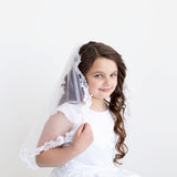 A young girl in a white dress with a veil stands against a plain white background, smiling at the camera.