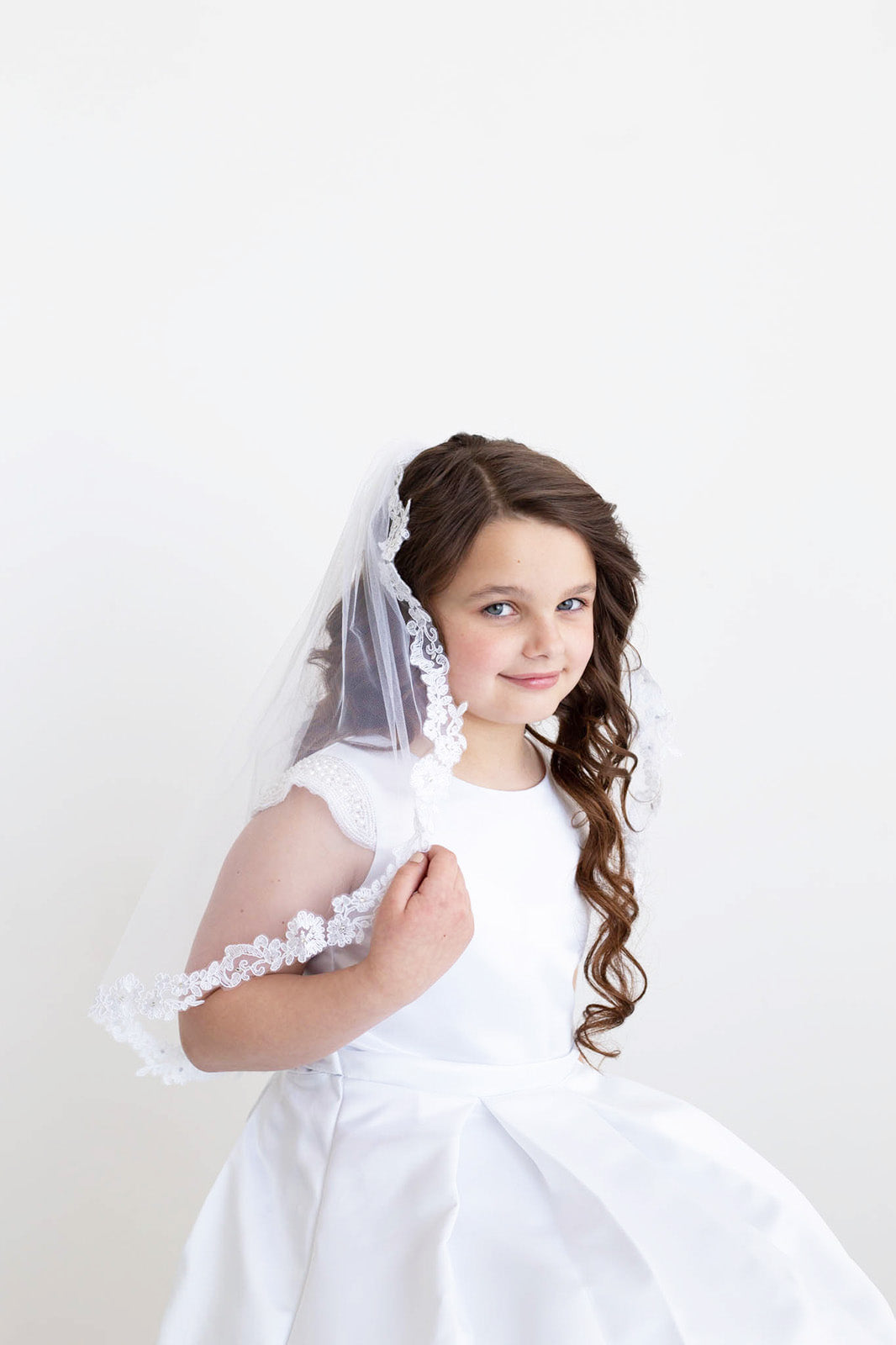 A young girl in a white dress with a veil stands against a plain white background, smiling at the camera.