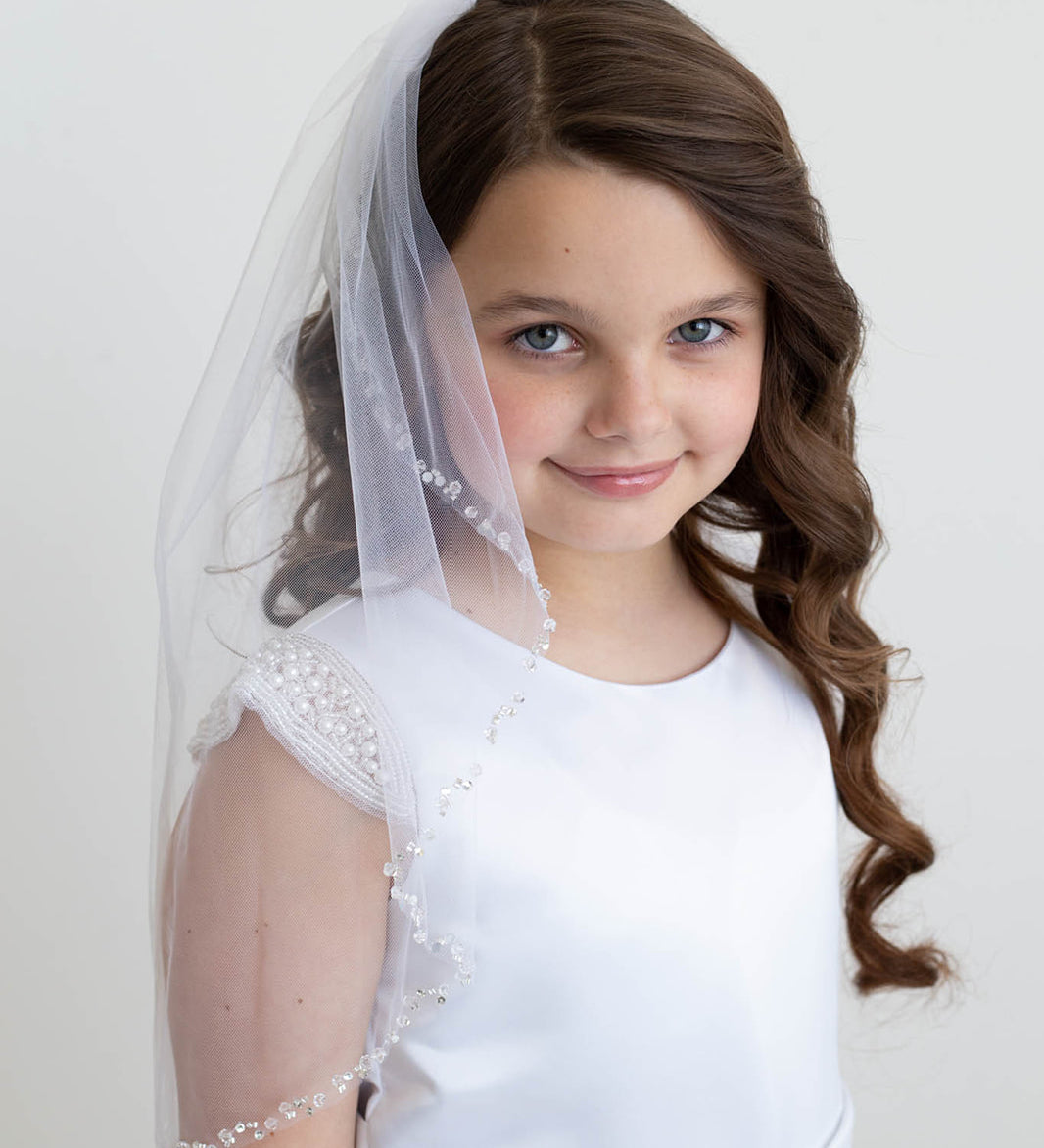 A young girl with curly brown hair wearing a white dress and veil, smiling at the camera.