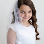 A young girl with curly brown hair wearing a white dress and veil, smiling at the camera.