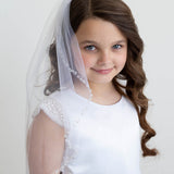 A young girl with curly brown hair wearing a white dress and veil, smiling at the camera.
