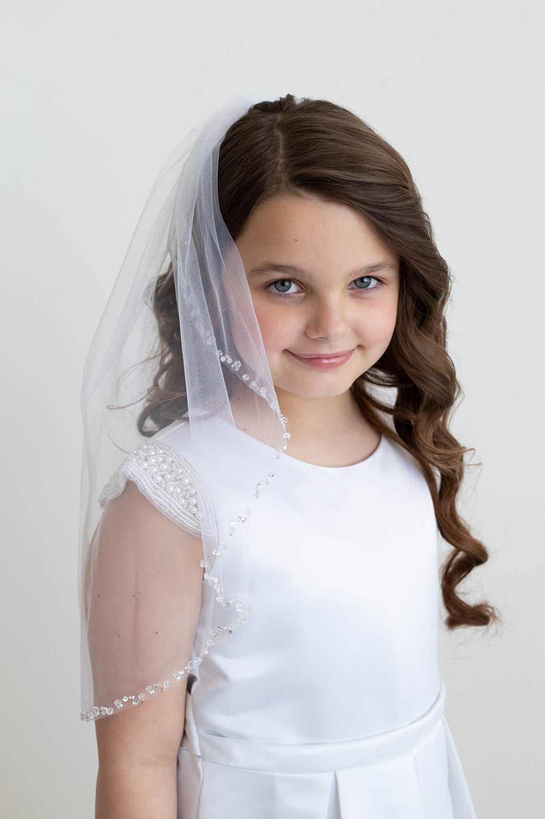 A young girl with curly brown hair wearing a white dress and veil, smiling at the camera.