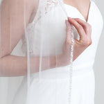 A close-up of a person wearing a white wedding dress with a veil, holding the veil with their hand.