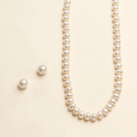 A necklace with a row of white pearls and two matching earrings are displayed against a light beige background.