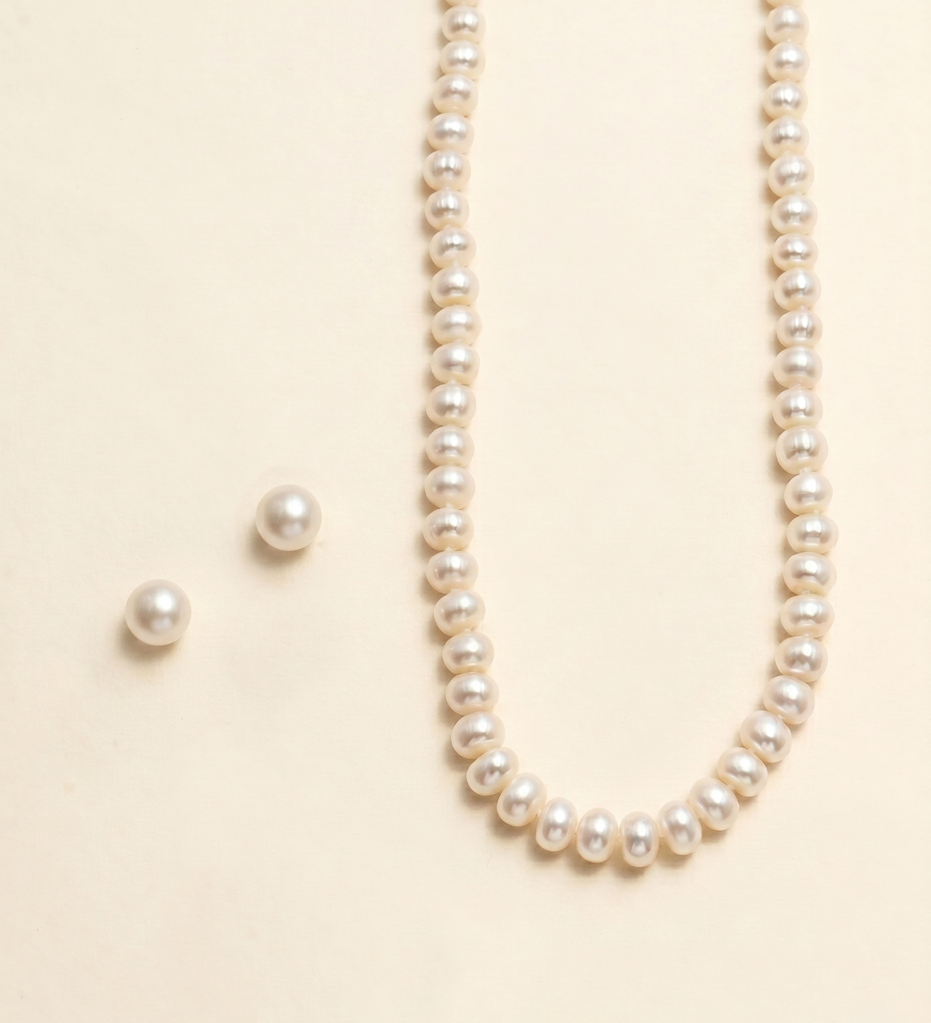 A necklace with a row of white pearls and two matching earrings are displayed against a light beige background.
