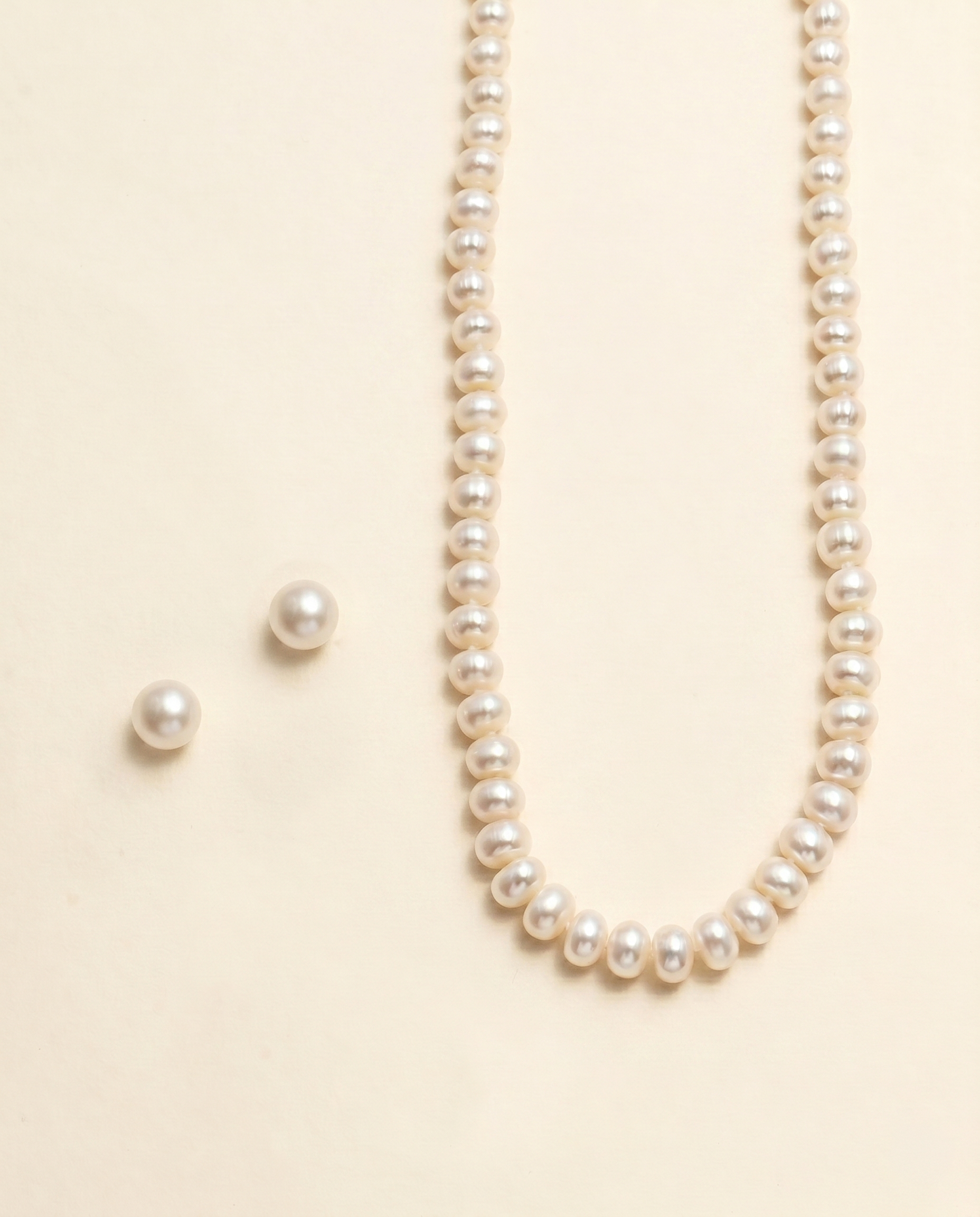 A necklace with a row of white pearls and two matching earrings are displayed against a light beige background.