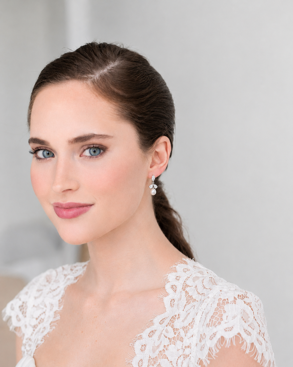 A woman with brown hair and blue eyes wearing a white lace dress and earrings, smiling at the camera.