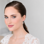 A woman with brown hair and blue eyes wearing a white lace dress and earrings, smiling at the camera.