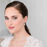 A woman with brown hair and blue eyes wearing a white lace dress and earrings, smiling at the camera.