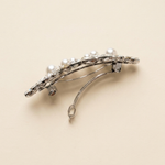 A silver hair clip adorned with pearls and diamonds is shown against a light beige background.