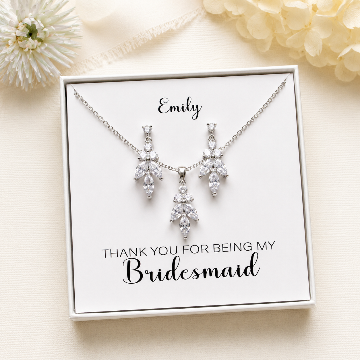 A necklace and earrings set with a personalized message on a white card, placed on a beige background with a white flower.