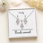 A necklace and earrings set with a personalized message on a white card, placed on a beige background with a white flower.