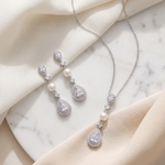A necklace and earrings with pearls and diamonds are displayed on a marble surface, with a beige cloth draped over the background.
