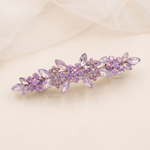 A decorative hair clip with purple flowers and gemstones is displayed on a light-colored surface.